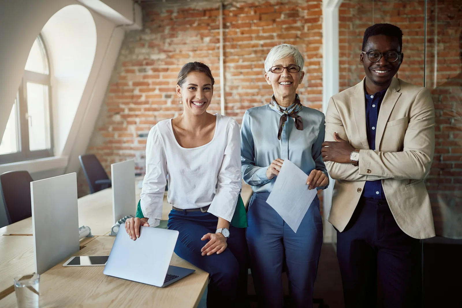 Drei lächelnde Kollegen, zwei Frauen und ein Mann, stehen in einem Büro mit Ziegelwänden und Laptops.